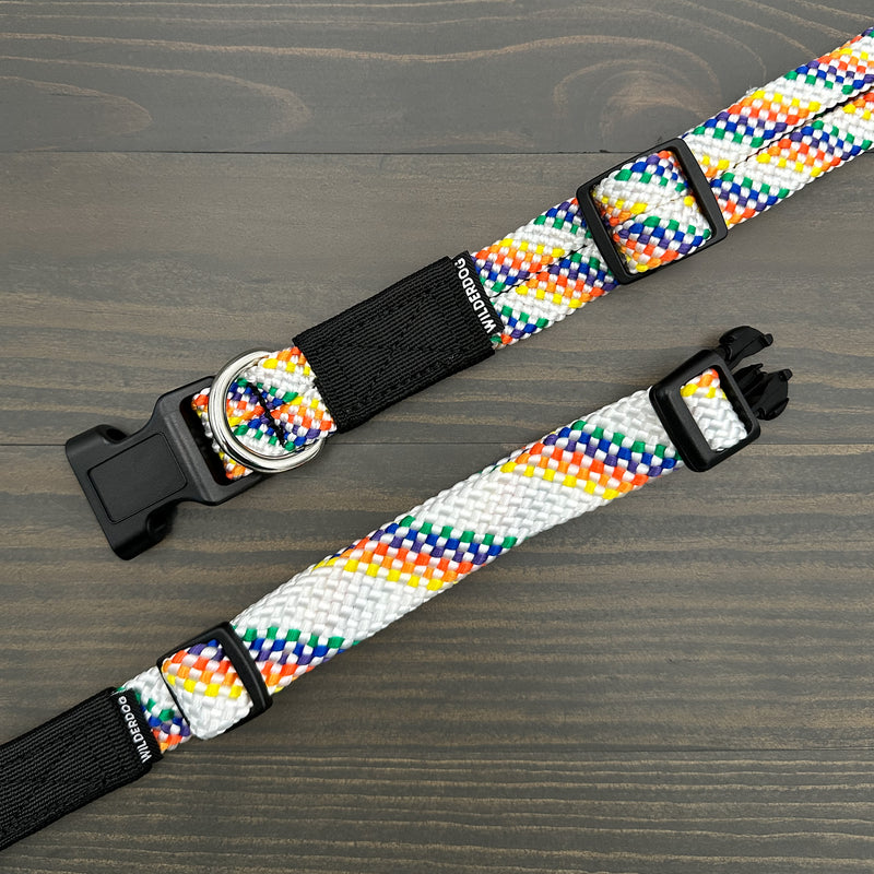 Displayed on a wooden surface are two Rainbow Collars, showcasing vibrant and braided patterns. Each heavy-duty collar features a black plastic buckle, a metal D-ring, and an eye-catching design. Both collars come with black tags that have white text, underscoring their durability and lifetime warranty.